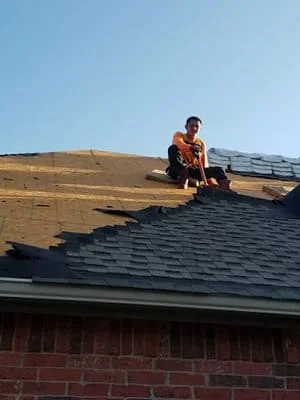 Roofer installing dark asphalt shingles on residential roof with brick home below